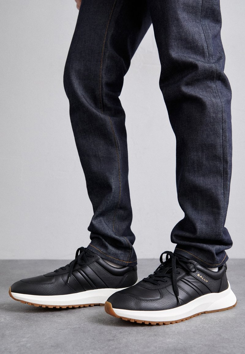 Zapatillas negras de cuero con un acabado texturizado, media suela blanca y suela de goma marrón; cuentan con cordones y tres franjas horizontales.