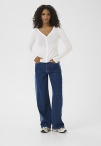 Vit, ribbad kofta med V-ringning och knappstängning, matchad med vida, mörkblå jeans. Modellen har på sig sneakers.