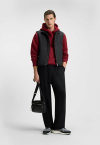 Chaleco negro acolchado sobre una sudadera con capucha roja, pantalones negros de pierna ancha y zapatillas grises. Lleva un bolso pequeño negro con cremalleras. Diseño minimalista.