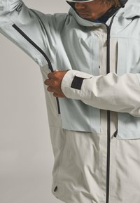 Light blue and cream waterproof jacket with black zippers, velcro accents, a hood, and a textured finish. Features adjustable cuffs and pockets.