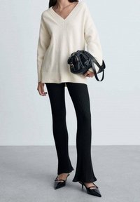 Pull en tricot crème avec col en V, pantalon flammé noir à côtes, et sac à main noir. Les chaussures présentent un bout pointu avec de petites décorations.