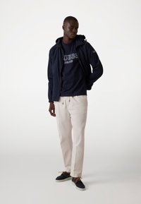 Navy blauwe lichtgewicht jas met capuchon, gedragen over een navy graphic t-shirt. Gecombineerd met crèmekleurige broek en donkere schoenen met bruine veters.