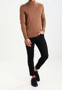 Jersey de cuello alto marrón hecho de un material suave, combinado con pantalones ajustados negros que presentan detalles de lado con cordones y zapatos negros.