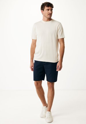 Cream short-sleeve shirt paired with dark navy shorts and white sneakers. The shirt has a smooth texture and a classic crew neckline.