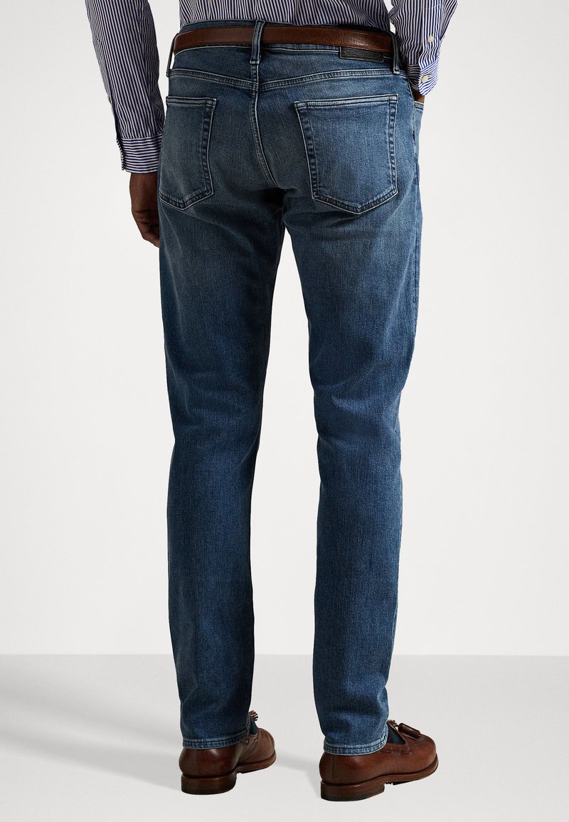 Blue denim jeans with a straight fit, featuring two back pockets and a mid-rise waist, worn with a brown leather belt and shoes.