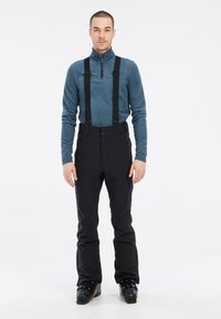 Zwarte skibroeken met verstelbare bretels, met een soepele textuur en een aansluitend ontwerp. Gedragen boven een blauwe fleece met lange mouwen.