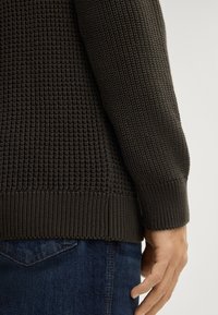 Donkergroene gebreide trui met een gestructureerd oppervlak, geribde zoom en manchetten; te zien met een hand rustend op blauwe spijkerbroek.