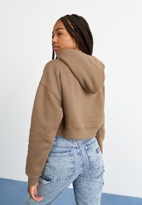 Hoodie corto marrone con cappuccio, indossato sopra jeans a vita alta in denim chiaro. Presenta un tessuto testurizzato e polsini a costine.