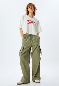 Gekürztes weißes T-Shirt mit rotem Text, kombiniert mit lockeren, olivgrünen Cargo-Hosen mit Seitentaschen und elastischem Bund. Legere Schuhe runden den Look ab.