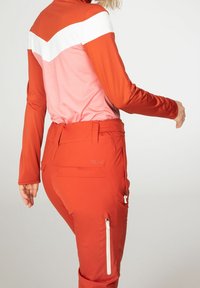 Tenue de ski masculine comprenant un haut à manches longues avec un design en chevrons rouge, rose et blanc, et un pantalon de ski orange avec des poches zippées.