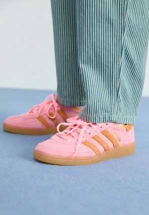 Personne portant un pantalon rayé turquoise et blanc et des baskets en daim rose avec des bandes beige, debout sur un sol bleu.