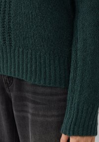 Dunkelgrüner, gestrickter Pullover mit Manschetten und Saum über schwarzen Jeans mit sichtbarer Hand an der Seite.