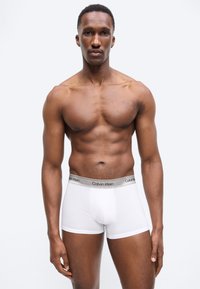 Homme musclé debout face à l'avant, portant un boxer blanc Calvin Klein sur un fond clair uni.