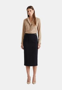 Blusa beige con bottoni e due tasche frontali, abbinata a una gonna a matita nera aderente con cuciture verticali e un dettaglio in tono oro.