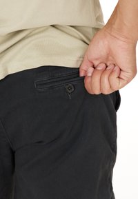 Svarta shorts med en knäppt bakficka. Tyget verkar vara strukturerat, och shortsen är kombinerade med en ljus beige skjorta.