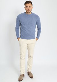 Man staat met zijn handen in zijn zakken, gekleed in een blauwe trui, beige broek en bruine suède laarzen tegen een effen witte achtergrond.