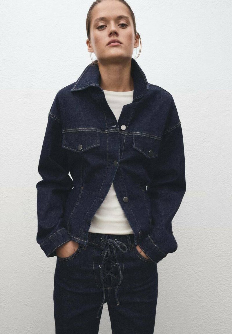 Chaqueta de mezclilla oscura con un cuello estructurado, bolsillos frontales y costuras visibles. Combinada con pantalones de mezclilla oscuros que tienen una cintura con cordones.