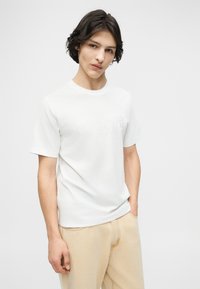 Νέος άνδρας με μεσαίου μήκους σκούρα μαλλιά, φορώντας απλό λευκό μπλουζάκι και μπεζ παντελόνι, σε λευκό φόντο.