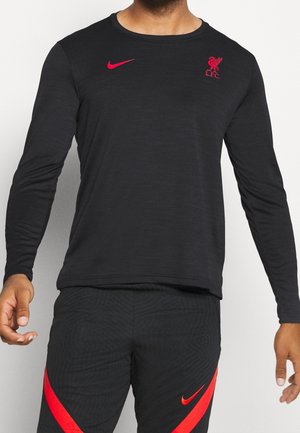 Mężczyzna ubrany w czarną bluzkę z długim rękawem i czarne spodnie z czerwonymi logo Nike i herbem Liverpool FC na piersi.