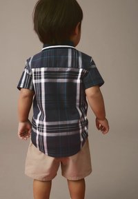 Camicia a maniche corte a quadri in blu navy con strisce bianche e verdi, abbinata a pantaloni corti beige. La vista posteriore mostra l'orlo arrotondato e i dettagli delle spalle.