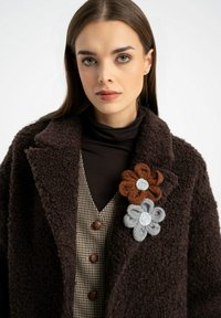 Brązowy futrzany płaszcz o teksturowanej powierzchni; nałożony na brązowy golf i kamizelkę w kratę. Trzy broszki w kształcie kwiatów w odcieniach brązu i szarości.