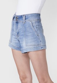 Šviesiai mėlynos džinsinės šortai su aukštu juosmeniu, dviem priekinėmis kišenėmis ir matomais siuvinėjimo detalėmis. Audinys turi šiek tiek nusidėvėjusį plovimą.