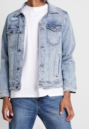 Mann trägt hellblaue Jeansjacke über weißem T-Shirt und mittelblauer Jeanshose, steht vor einfarbigem Hintergrund.