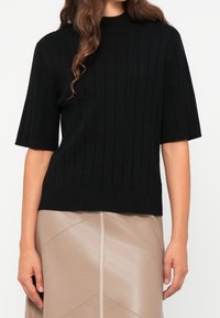 Haut noir en maille côtelée à manches courtes avec un col montant. Le tissu présente une texture lisse et un motif uniforme. Associé à une jupe beige.