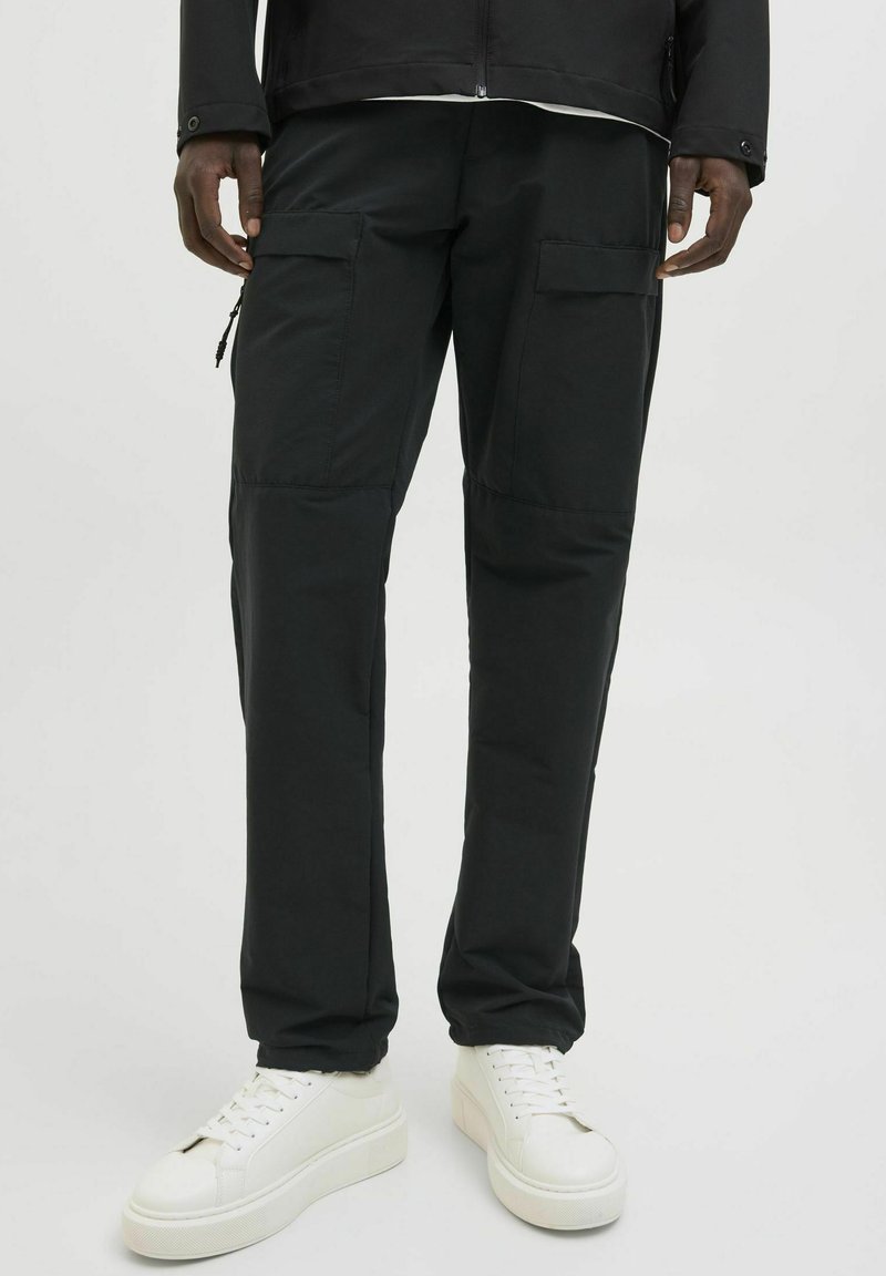 Schwarze Cargo-Hosen mit zwei großen Vorderstaschen, slim fit und glatter Textur. Kombiniert mit weißen Sneakers, was einen Farbkontrast schafft.