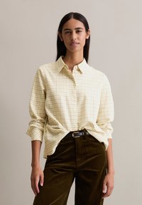 Button-up shirt in light yellow with a grid pattern; long sleeves rolled at the cuffs. Paired with dark green corduroy pants.