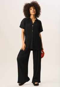 Black, pleated two-piece outfit featuring a button-up shirt with short sleeves and wide-leg pants; includes a red clutch and platform sandals.