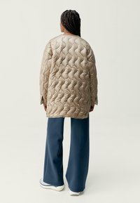 Béžová prošívaná bunda s texturovaným vzorem, volný střih a dlouhé rukávy, spárovaná s širokými tmavomodrými kalhotami a bílou obuví.