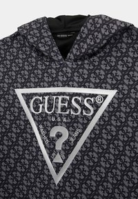 Sudadera negra con capucha que presenta un patrón de logotipos repetidos, un triángulo plateado prominente con el texto "GUESS®" y un gráfico de un signo de interrogación.