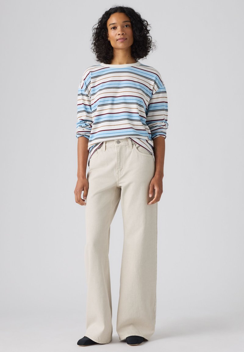 Femme aux cheveux bouclés portant une chemise à manches longues rayée bleu, blanc et marron et un pantalon ample beige clair, debout devant un fond uni.