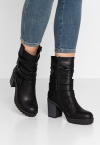 Botas de cuero negro hasta el tobillo con un tacón grueso, que presentan detalles de correas y una cremallera lateral. Textura suave con una suela con relieve para mayor agarre.