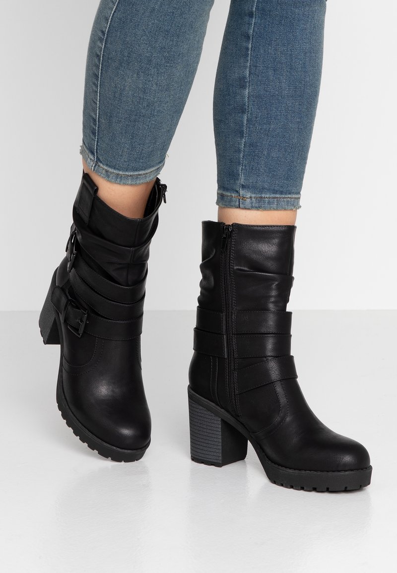 Botas de cuero negro hasta el tobillo con un tacón grueso, que presentan detalles de correas y una cremallera lateral. Textura suave con una suela con relieve para mayor agarre.