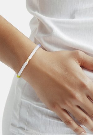 Handgelenk mit einem schlanken weißen Perlenarmband mit kleinen goldenen und pastellfarbenen Perlen, das auf einem weißen strukturierten Stoffkleidungsstück liegt.