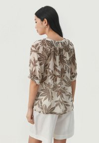 Gennemsigtig beige bluse med et bladmønster i brun og rust, med korte puffærmer og en samlet rygdetalje. Bæres med hvide shorts.