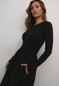 Mustat pitkähihaiset mekko, jossa on tiukasti istuva ribbitoppi ja virtaava, kerrostettu hame. Sisältää sivutaskut ja pyöreän pääntien.