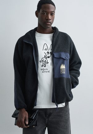 Zwarte fleec jas met marineblauwe accenten, rits aan de voorkant, twee zakken, met een logo patch. Draag over een witte grafische t-shirt. Donkergrijze spijkerbroek.