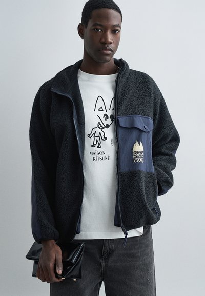 Schwarze Fleecejacke mit navyfarbenen Akzenten, Reißverschluss vorne, zwei Taschen, mit einem Logo-Patch. Getragen über einem weißen Grafik-T-Shirt. Dunkelgraue Jeans.