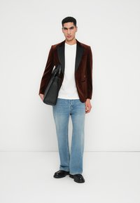 Bruine fluwelen blazer met zwarte revers, witte t-shirt en lichtblauwe wijde spijkerbroek. Model houdt een zwarte tote bag vast en draagt zwarte schoenen.
