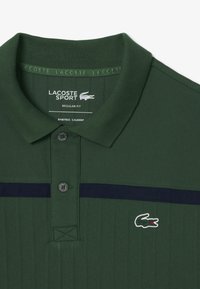 Grön polotröja med texturerat tyg, en marinblå rand, en tvåknäppt knäppning och en liten vit och grön krokodil-logotyp.