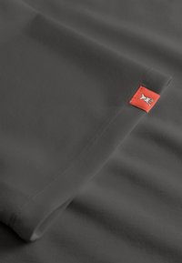 Donkergrijze stof met een omgeslagen rand en een klein oranje label met een wit hand symbool dat dicht bij de naad is gestikt.