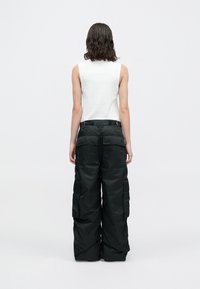 Persoon met schouderlang golvend haar, gekleed in een mouwloos wit topje en zwarte wijde cargobroek, die met de rug naar de camera staat op een eenvoudige achtergrond.