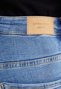 Gaiši zilu denim audumu bikses ar tan krāsas ādas plāksni, uz kuras ir norakstīts "ideālās bikses", un redzamām šuvju detaļām jostasvietā.