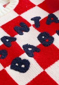 Červeno-bíle kostkovaný pletený materiál s vyvýšenými tmavě modrými texturovanými písmeny, která hlásají slovo "BANANA" a část slova "ANT" je viditelná.