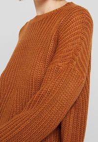 Maglione lavorato a maglia in un ricco colore marrone con un motivo testurizzato a coste. Presenta un collo a giro e maniche lunghe, mettendo in risalto un tessuto morbido.