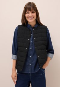 Donna sorridente che indossa un gilet trapuntato nero sopra una camicia di denim azzurro con le maniche arrotolate e jeans blu scuro.