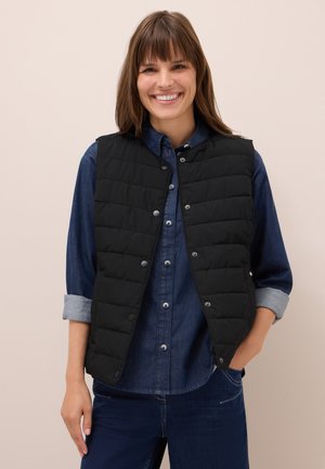 Donna sorridente che indossa un gilet trapuntato nero sopra una camicia di denim azzurro con le maniche arrotolate e jeans blu scuro.